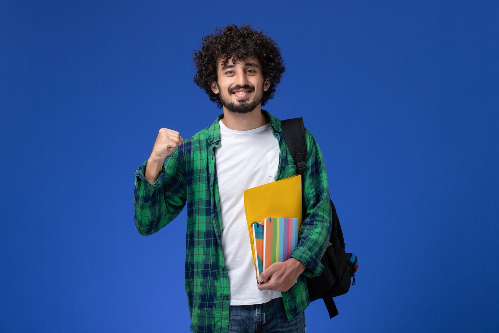 front view male student wearing black backpack holding copybooks files blue wall